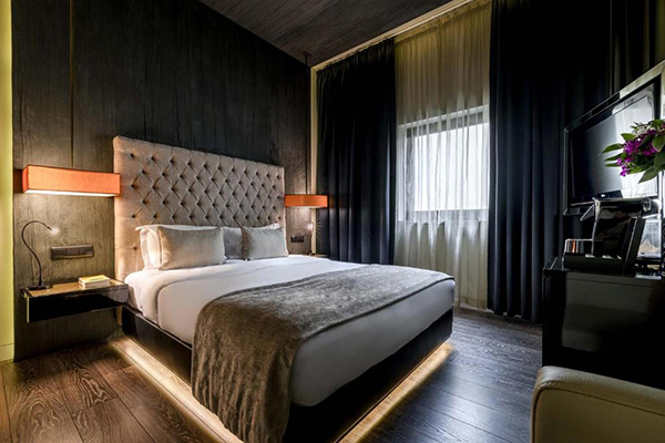 Um quarto bem escuro, com a imensa cama com cabeceira cinza e luminárias bem modernas laranjas