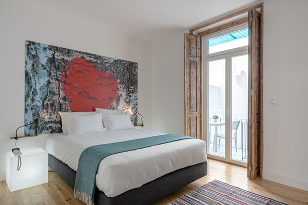 Um quarto bem claro, com uma pintura moderna e colorida em cima da cama. Tem uma varada com uma mesinha e cadeiras