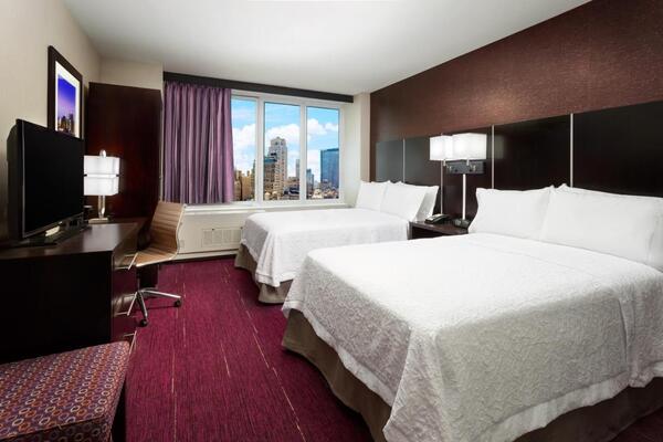 Quarto de hotel cuja decoração é nas cores marrom, branca e rosa. Duas camas de casal, TV, abajur, quadro e janela com vista para Nova York fazem parte da cena.