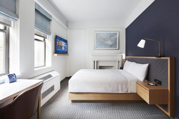 Quarto majoritariamente branco, com uma cama, luminária, TV, mesa e cadeira. Atrás da cama há uma parede na cor azul escuro.