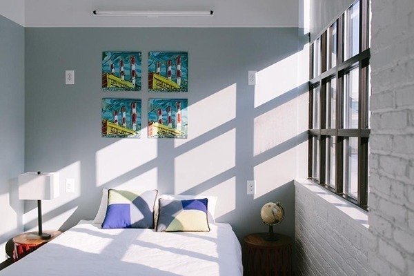 Um quarto de hotel com uma cama de casal com quadros acima da cama com desenhos coloridos, e a luz entra bem bonita através da janela