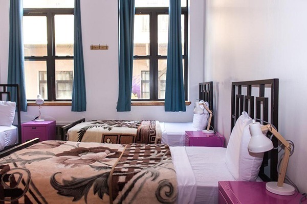 Um quarto de hostel com 3 camas com lençóis brancos e cobertos marrom, mesa de cabeceira rosa com luminária