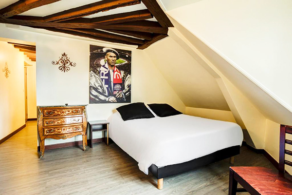 Quarto com cama de casal branca com travesseiros pretos, teto baixo, cômoda antiga, teto com madeira e paredes brancas