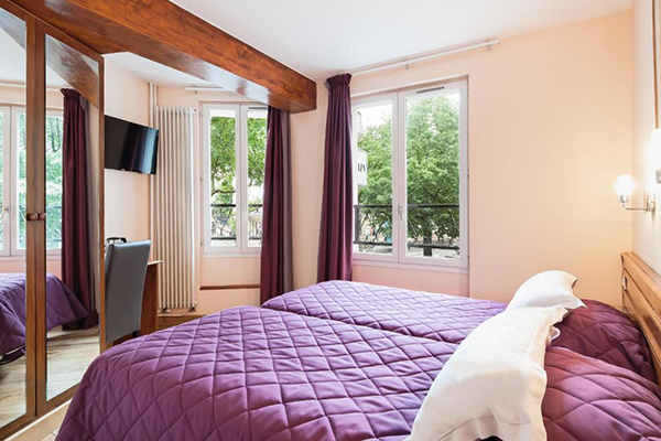 Quarto de hotel amplo com 2 janelas grandes, 2 camas de solteiro juntas com roupa de cama roxa, ármario com espelho