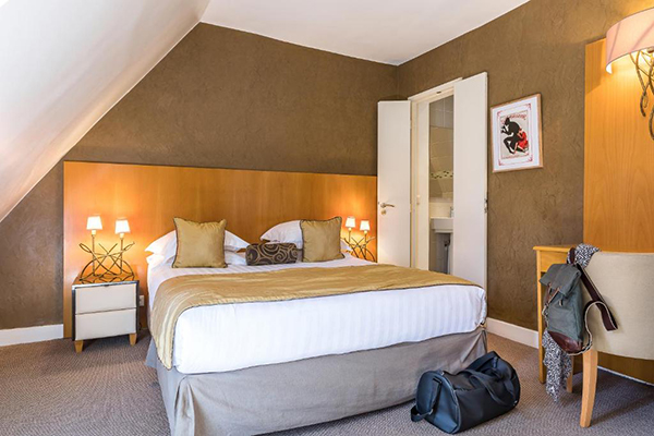 Um quarto de hotel com uma cama de casal grande branca com detalhes dourados. Cabeceira de madeira clara e papel de parede marrom-dourado