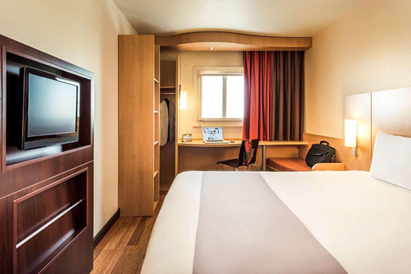 Um quarto de hotel com janela pequena, cama de casal branca, tv e escrivaninha com um laptop 