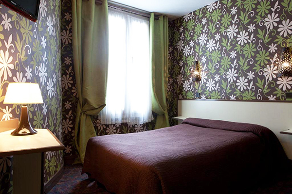 Quarto de hotel com papel de parede escuro com flores verdes e brancas, cama com roupa de cama escura e janela