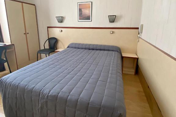 Quarto de hotel claro, com cama com roupa de cama cinza e ármario claro