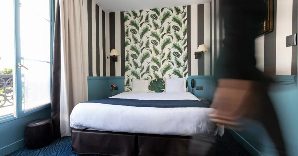 Hotéis Baratos em Paris: um quarto de hotel com papel de parede colorido e cama branca, com uma pessoa passando do lado direito
