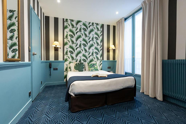 Quarto de hotel com paprel de parede verde, paredes azuis, cama de casal branca com detalhes tropicais verdes e azuis