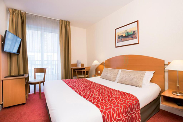 Quarto de hotel com cama de casal branca comd etalhes vermelhos, cabeceira de madeira, escrivaninha e tv