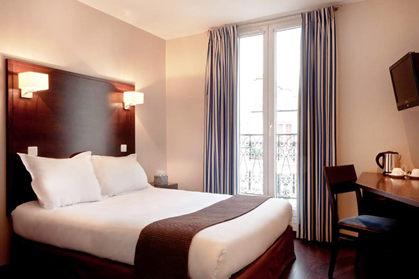 Quarto de hotel com cama de casal branca, cabeceira de madeira escura, janela grande e escrivaninha
