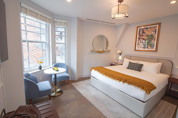 Um quarto com uma cama giganre com detalhes em amarelo, com paredes claras e janelas grandes. Duas poltronas azuis e uma mesinha redonda