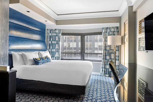Um quarto de hotel azul, com cabeceira acolchoada, cama branca, carpete e cortinas com desenhos azuis