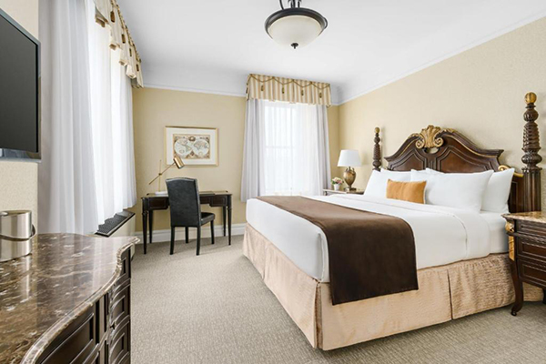 Quarto de hotel com cama alta em estilo antigo, detalhes em marrom, cortinas brancas