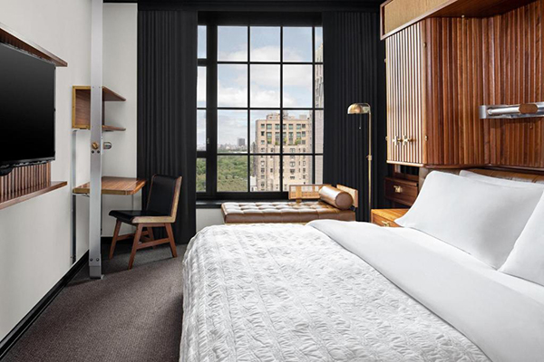 Quarto de hotel com paredes brancas, móveis em madeira, janela enorme com vista para o Central Park, cortinas pretas e divã ao lado da janela