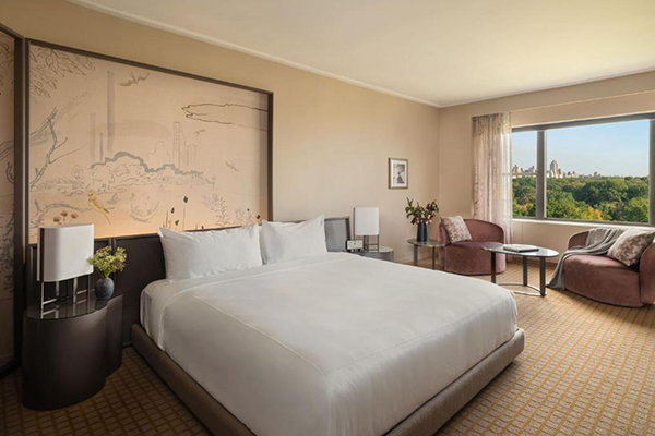 Um quarto de hotel comprido, com paredes bege, cama grande branca, uma pintura oriental na parede e vista do Central Park da janela