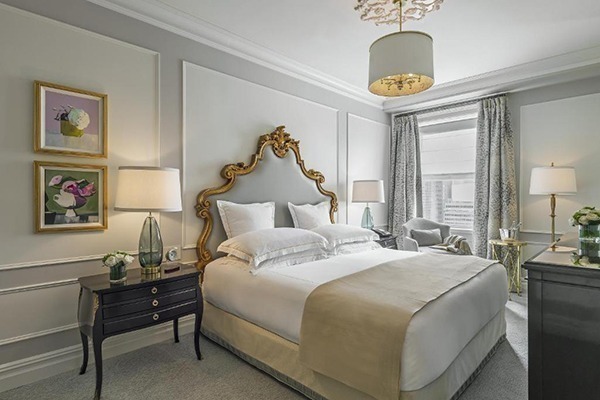 Quarto de hotel com cama luxuosa em tons cinza e dourado, paredes brancas e cinzas