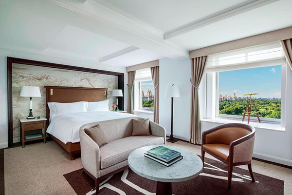 Quarto de hotel imenso, com duas janelas com vista para o Central Park, cama grande branca, sofá e poltrona em volta de uma mesa redonda
