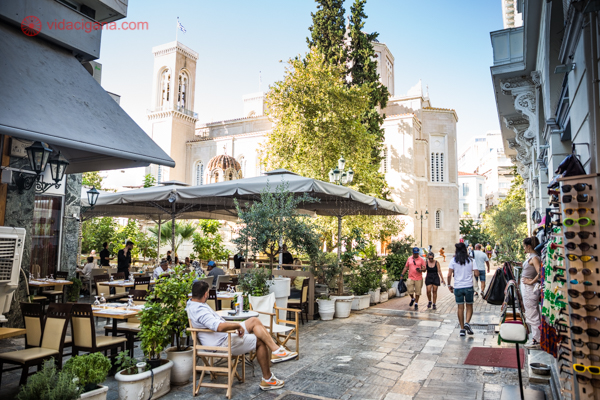 Uma rua em Atenas cheia de cafés e restaurantes, com pessoas sentadas e outras caminhando, uma igreja ao fundo e árvores