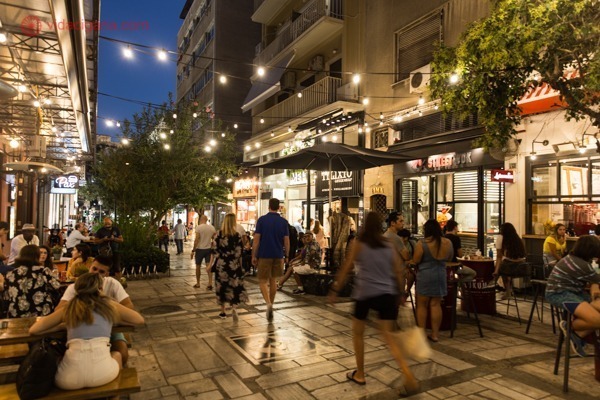 Uma rua bem movimentada de Atenas, com muitos restaurantes, mesas, pessoas sentadas e outras caminhando. Muitas luzes