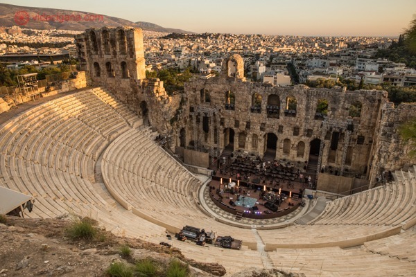 Um teatro romano em meia lua, com um palco lá embaixo e arcos em seu fundo, tudo muito bem conservado. A cidade ded Atenas se espalha ao fundo