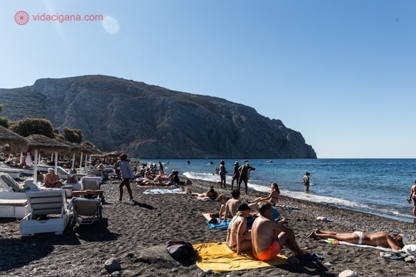 Uma praia em Santorini, com muitas pessoas na areia pegando sol, espreguiçadeiras e guarda-sóis do lado esquerdo e um monte alto ao fundo