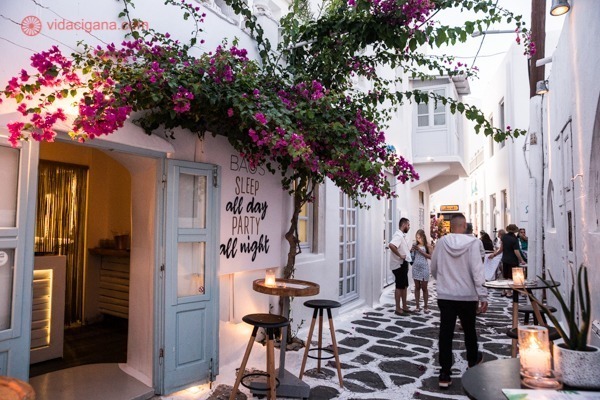 Melhor época para visitar a Grécia: Uma rua toda branca em Mykonos, com flores rosas acima da porta de um bar