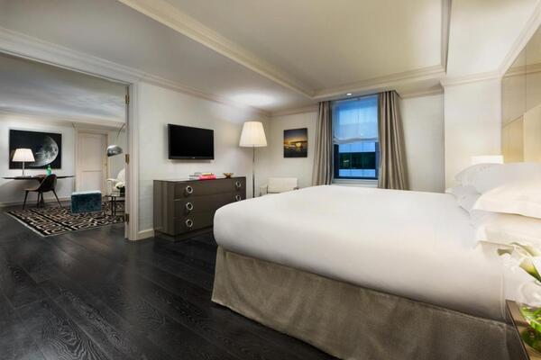 Quarto de hotel completo, dividido em dois ambientes. Conta com cama de casal, TV, cômoda e outros elementos.