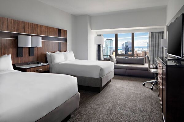Imagem de um quarto com duas camas de casal e um sofá-cama perto da janela, com vista para Nova York. Abajur, TV, cômoda e mesa de escritório também fazem parte da cena.