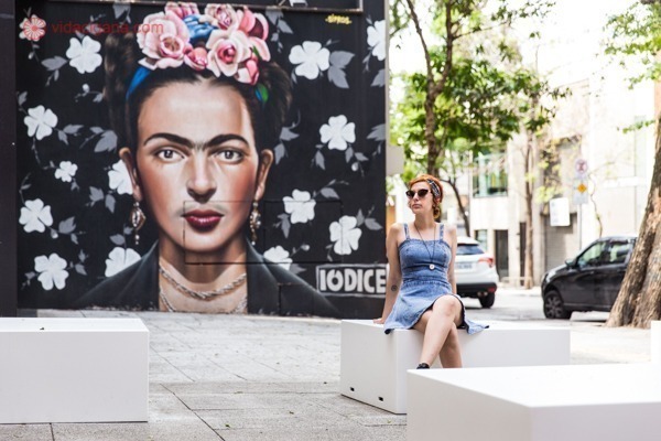Uma mulher sentada num banco branco com um mural da Frida Khalo atrás