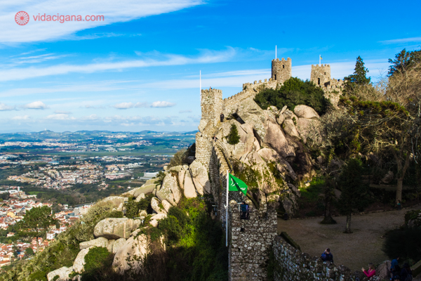 O Castelo dos Mouros com suas muralhas em pedra circulando as montanhas bem alto, com uma bandeira verde tremulando e a região toda abaixo