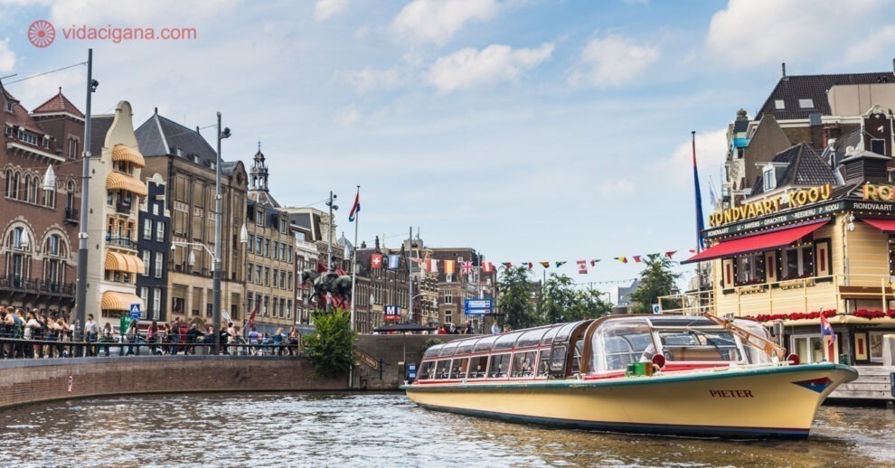 Melhores passeios de barco em Amsterdam: Um barco comprido com o teto de vidro navegando por um dos canais de Amsterdam, com vários prédios ao redor