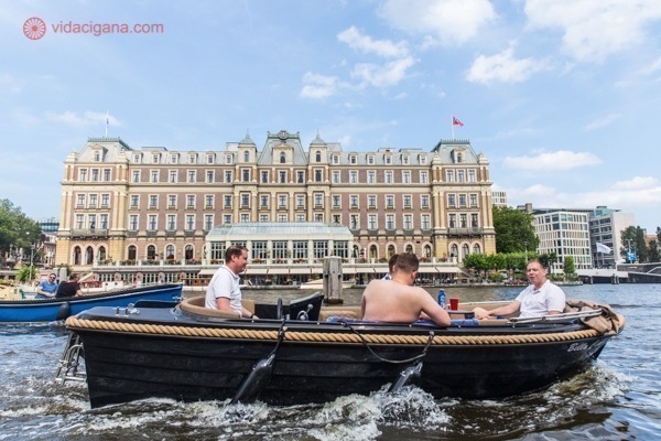 Quatro homens dentro de um barco privado passando por prédios históricos de Amsterdam num dia de sol