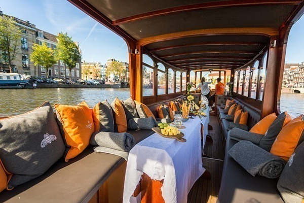 O interior de um barco comprido com uma mesa bem comprida com queijos e vinhos, e bancos nas laterais do barco com almofadas coloridas