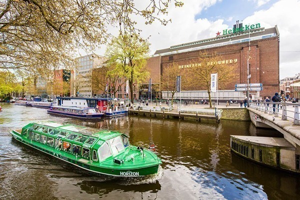 Um barco comprido verde passando em frente ao prédio onde acontece o Heineken Experience