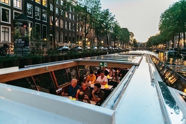 Várias pessoas dentro de um barco comprido durante o entardecer, passando por um dos canais de Amsterdam