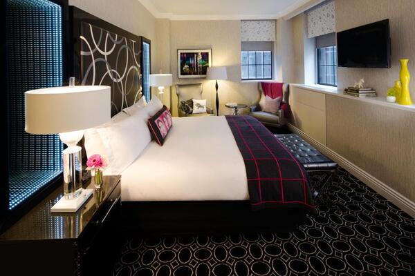 Imagem de quarto de hotel, com piso preto com estampas circulares. O restante do quarto é nas cores branco e reto e conta com itens em vermelho.