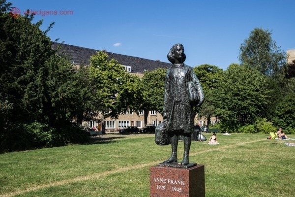 A estátua de Anne Frank segurando duas pastas nos braços num lindo jardim cheio de verde num dia de sol
