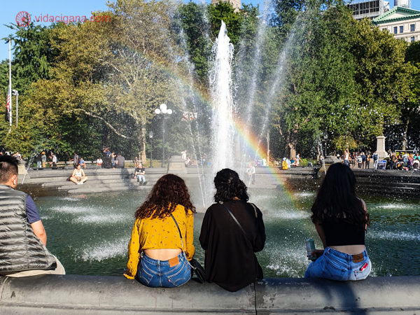Três pessoas sentadas na fonte de Greenwich Village Park com um arco íris na água 