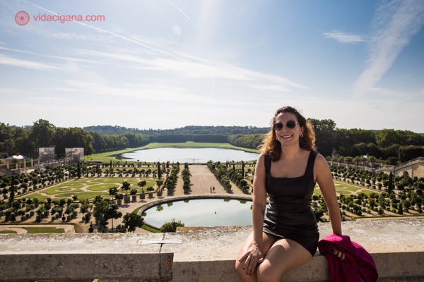 Eu sentada em uma mureta de frente aos jardins do Palácio de Versailles