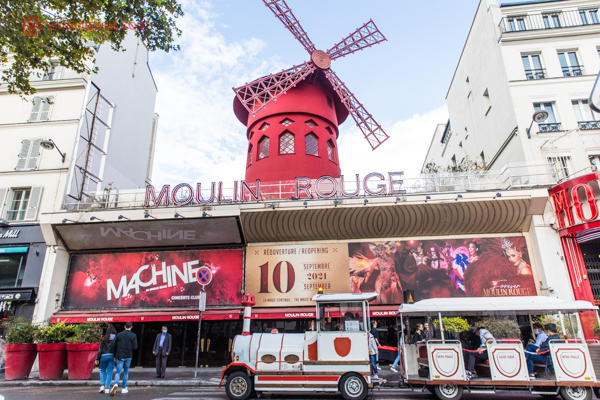 A fachada do Moulin Rouge com seu moinho vermelho e um trenzinho branco parado bem a frente