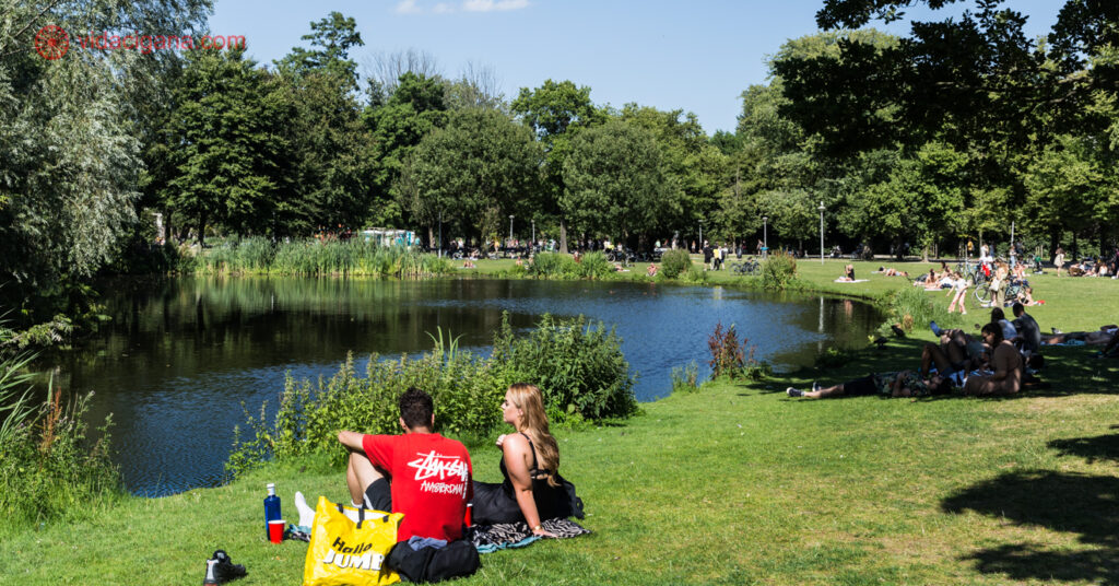 Pessoas sentandas no gramado em um parque em Amsterdam ao redor de um lago num dia de sol