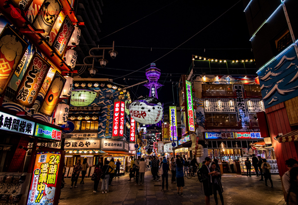 Imagem noturna do distrito de Shinsekai em Osaka, com suas luzes de neon e a torre Tsutenkaku iluminada ao fundo. Pessoas passeiam entre as coloridas fachadas de lojas e restaurantes tradicionais japoneses.




