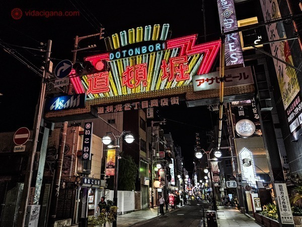 A fotografia mostra a entrada do distrito de Dotonbori em Osaka, caracterizada pelo icônico letreiro neon que anuncia "Dotonbori". Abaixo do letreiro, a rua é iluminada por luzes suaves e repleta de várias lojas e restaurantes de ambos os lados, com pessoas caminhando e explorando a área. 