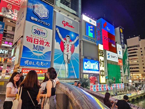 Distrito de Dotonbori em Osaka à noite, com um grupo de pessoas à beira de um canal. Grandes painéis publicitários e letreiros iluminados cobrem as fachadas dos edifícios, destacando-se uma figura animada de um corredor. O ambiente é urbano e vibrante, refletindo a vida noturna japonesa.




