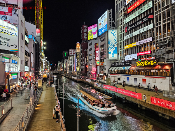 O que fazer em Osaka: Ccanal de Dotonbori em Osaka, Japão, à noite. Um barco cheio de passageiros navega pelo canal, enquanto pedestres passeiam pelo calçadão iluminado. As margens do canal são decoradas com uma miríade de anúncios luminosos e letreiros em japonês, refletindo a energia vibrante e a cultura pop do distrito.
