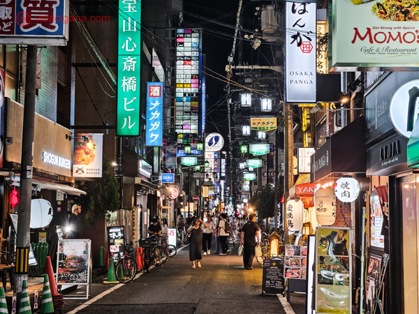 A imagem captura uma rua estreita e movimentada em Osaka, Japão, à noite. Letreiros em japonês e iluminação neon adornam as fachadas dos edifícios, enquanto pedestres passeiam e exploram a variedade de restaurantes e lojas locais. Bicicletas estão estacionadas ao lado da calçada, e a cena transmite uma sensação acolhedora e íntima da vida urbana japonesa.




