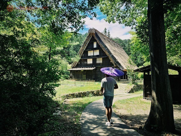 Carlos andando com uma sombrinha japonesa roxa em direção à uma casa antiquíssima japonesa com o teto feito de palha