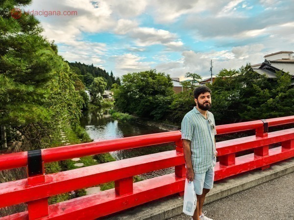 Carlos parado olhando pra câmera na Ponte Nakabashi, uma ponte vermelha com o rio a passar embaixo cercado de verde e com montanhas ao fundo